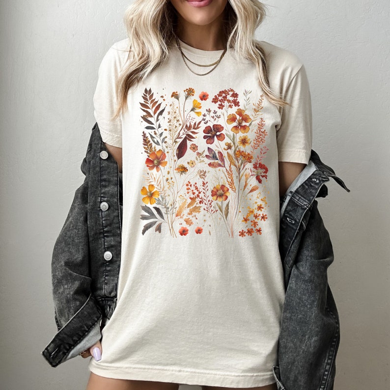 Puede incluir: Una camiseta beige con un estampado floral que presenta una variedad de flores en tonos naranja, rojo y amarillo. Las flores est&aacute;n dispuestas en un racimo en la parte delantera de la camiseta.