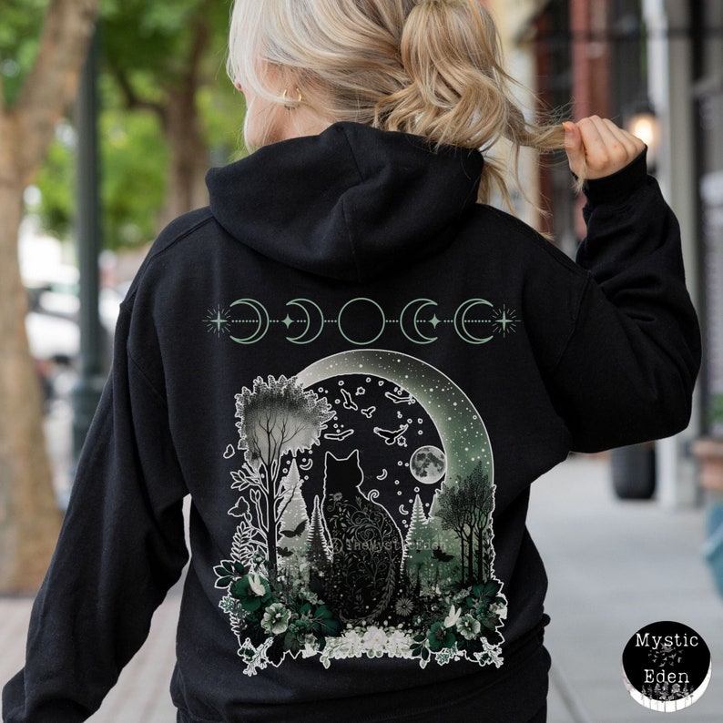 May include: Black hooded sweatshirt with a green and white graphic print of a cat sitting in a forest with a crescent moon and stars. The graphic also includes the text "Mystic Eden".