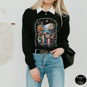K&ouml;nnte beinhalten: Schwarzes Sweatshirt mit einem Aufdruck einer B&uuml;cherstapel, umgeben von Blumen und einem Zitat, das lautet: "Ein Buch ist eine magische Sache, die es dir erm&ouml;glicht, an ferne Orte zu reisen, ohne jemals zu gehen."