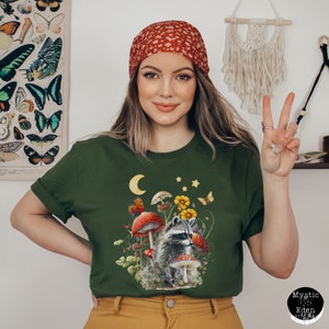 May include: A green t-shirt with a graphic of a raccoon standing in a forest scene with mushrooms, flowers, and a crescent moon. The raccoon is gray and black with a white mask. The mushrooms are red with white spots. The flowers are yellow and orange. The moon is white. The stars are yellow.