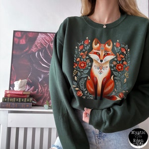 Op de afbeelding: Een donkergroen sweatshirt met een kleurrijke illustratie van een vos die in een krans van bloemen zit. De vos is oranje, rood en wit met een bloemenkraag. De bloemen zijn rood, oranje, geel en groen.