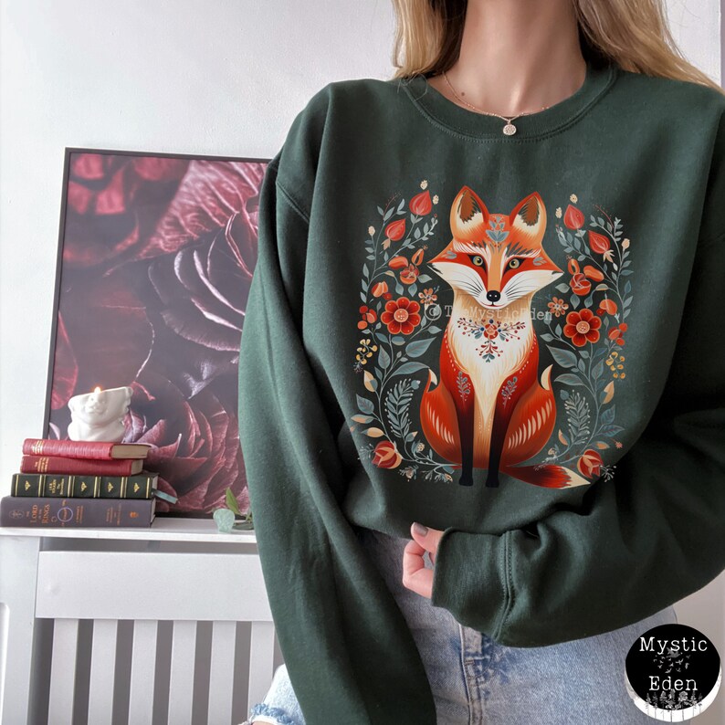 Op de afbeelding: Een donkergroen sweatshirt met een kleurrijke illustratie van een vos die in een krans van bloemen zit. De vos is oranje, rood en wit met een bloemenkraag. De bloemen zijn rood, oranje, geel en groen.