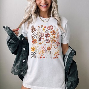 Puede incluir: Una camiseta blanca con un estampado floral con una variedad de flores y hojas de color naranja, rojo y amarillo.