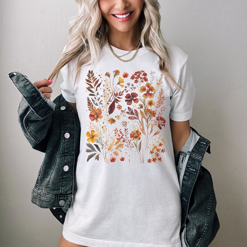 Puede incluir: Una camiseta blanca con un estampado floral con una variedad de flores y hojas de color naranja, rojo y amarillo.