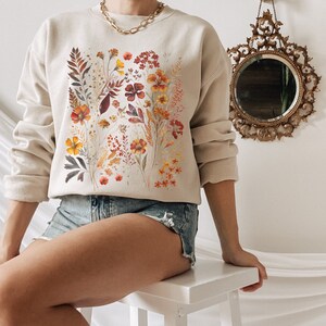 May include: A beige sweatshirt with a floral print featuring orange, yellow, and brown flowers. The flowers are arranged in a vertical pattern on the front of the sweatshirt.