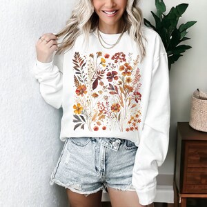 May include: White sweatshirt featuring a watercolor floral design in shades of orange, yellow, and brown. The botanical illustration includes various flowers and foliage, creating a nature-inspired aesthetic. The sweatshirt has long sleeves and a crew neck.