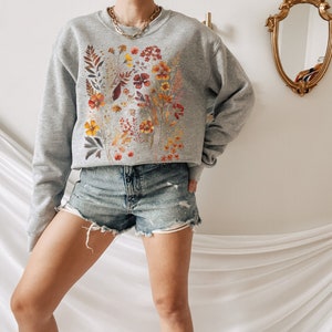 May include: A grey sweatshirt with a floral print featuring orange, yellow, and red flowers. The flowers are arranged in a vertical pattern on the front of the sweatshirt.
