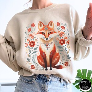 Op de afbeelding: Een beige sweatshirt met een kleurrijke illustratie van een vos die in een bloemenpatroon zit. De vos is oranje, rood en wit met een bloemenontwerp rond zijn nek. De bloemen zijn rood, oranje en geel.