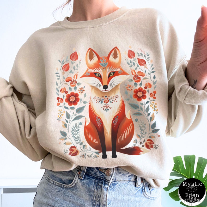 Op de afbeelding: Een beige sweatshirt met een kleurrijke illustratie van een vos die in een bloemenpatroon zit. De vos is oranje, rood en wit met een bloemenontwerp rond zijn nek. De bloemen zijn rood, oranje en geel.