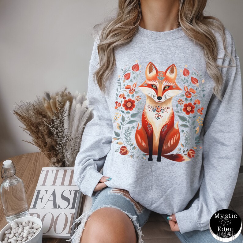 Op de afbeelding: Een grijs sweatshirt met een kleurrijke illustratie van een vos die in een bloemenkrans zit. De vos is oranje en rood met een witte borst en een zwarte neus. De bloemen zijn rood, oranje, geel en groen.