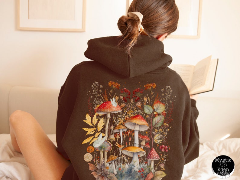 Op de afbeelding: Een bruine hoodie met een kleurrijk ontwerp van paddenstoelen en bloemen op de rug. Het ontwerp bevat rode, oranje, gele en groene paddenstoelen met witte stippen, omringd door groene bladeren en bloemen.