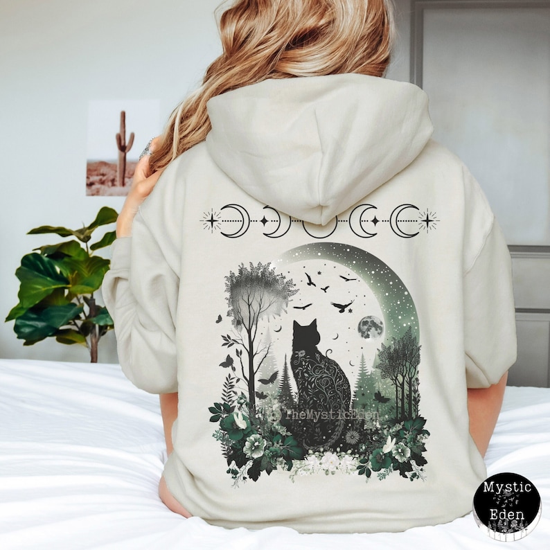 May include: A cream-colored hooded sweatshirt with a black cat silhouette sitting in a forest scene with a crescent moon and stars. The text "The Mystic Eden" is printed on the sweatshirt.