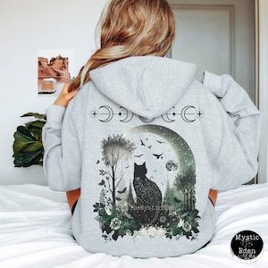 May include: Gray hooded sweatshirt with a black cat silhouette sitting in a moonlit forest scene. The design features a crescent moon, stars, and a variety of trees and flowers. The text "The Mystic Eden" is visible at the bottom of the design.