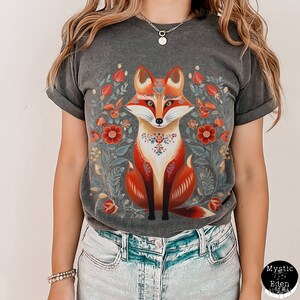 May include: Dark grey t-shirt with a colourful fox design and floral elements. The fox is rendered in orange, red, and white, set against a backdrop of flowers in a folk art style. The t-shirt has a relaxed fit and rolled sleeves.