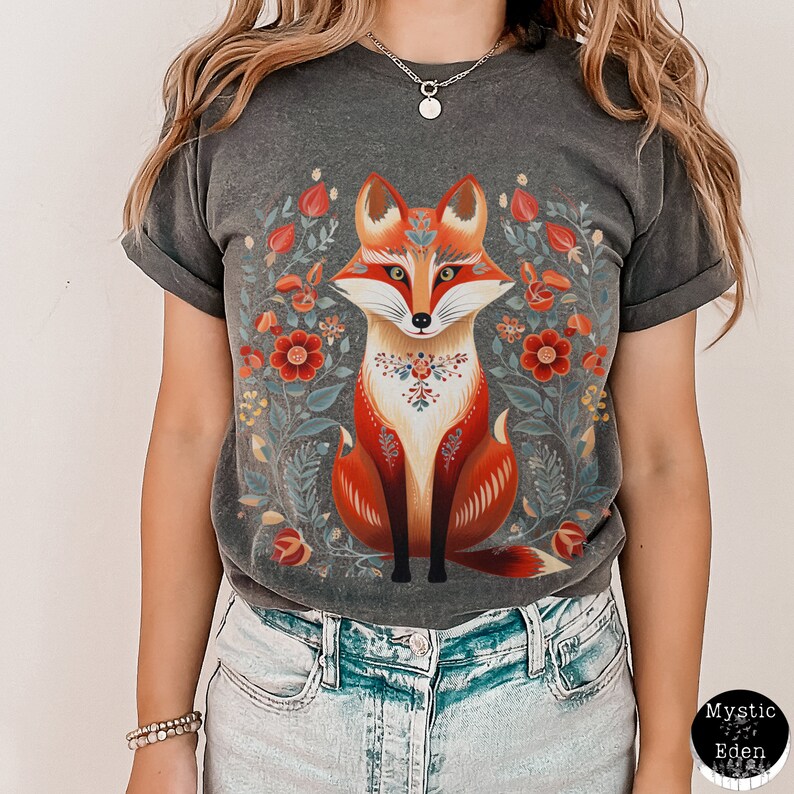 May include: Dark grey t-shirt with a colourful fox design and floral elements. The fox is rendered in orange, red, and white, set against a backdrop of flowers in a folk art style. The t-shirt has a relaxed fit and rolled sleeves.