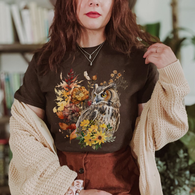 May include: A brown t-shirt with a graphic print of an owl surrounded by fall foliage and flowers. The owl is brown and white with yellow eyes. The flowers are yellow and orange.