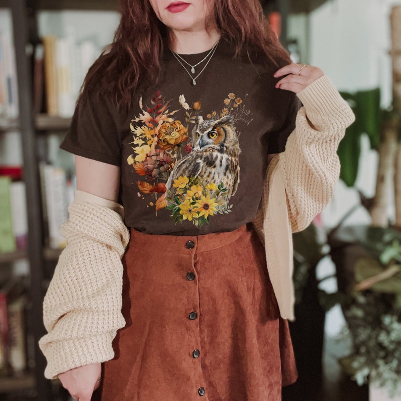 May include: A brown t-shirt with a graphic print of an owl surrounded by fall foliage. The owl is brown and white with yellow eyes. The foliage is in shades of brown, yellow, and orange. The shirt is being worn by a person with a brown skirt and a cream cardigan.