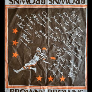 Puede incluir: Toalla del equipo de f&uacute;tbol americano Browns en marr&oacute;n y blanco con detalles en naranja. Presenta un gr&aacute;fico de un jugador de f&uacute;tbol, estrellas naranjas y firmas de jugadores. Las palabras "BROWNS" est&aacute;n impresas arriba y abajo.