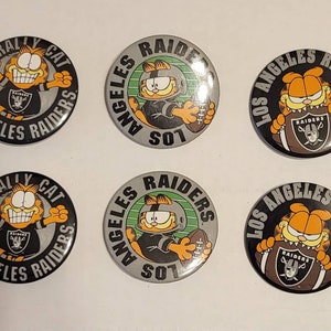 May include: Six round buttons featuring Garfield in Los Angeles Raiders attire. The buttons have a black, silver, and orange color scheme. Some buttons say "Rally Cat" and others say "Los Angeles Raiders".