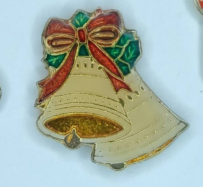 May include: Two gold-colored bells with a red and green holly and bow design. The bells are decorated with a gold glitter finish.