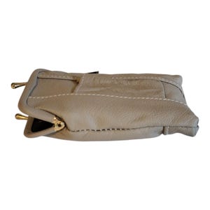 May include: A beige leather clutch bag with a flap closure and gold-toned hardware. The bag features white stitching and a rectangular shape. Designed for hand-carrying, it's suitable for various occasions. The bag is approximately 20 cm long.