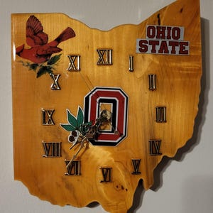Peut inclure: Horloge en bois en forme d'Ohio, avec un cardinal rouge, le logo de l'Ohio State et les mots "OHIO STATE". Elle comporte des chiffres romains et des aiguilles dorées. L'horloge est en bois de couleur claire.