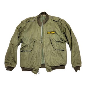 Op de afbeelding: Olijfgroene bomberjack met geribbelde kraag, manchetten en tailleband. Voorzien van twee voorzakken en een ritssluiting. De jas heeft een "U.S. ARMY" patch op de borst. Een vintage jas in militaire stijl.