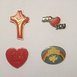 May include: Four enamel pins with religious themes. A red cross with a gold outline and a figure of a person with outstretched arms. A red heart with a gold outline and the words "GOD" and "YOU". A red heart with a gold outline and the words "JESUS LOVES ME". A blue oval with a rainbow and the word "JESUS" and a white dove.