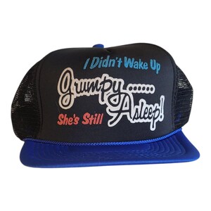 May include: Black and blue trucker hat with the text "I Didn't Wake Up Grumpy... She's Still Asleep!" in white, blue, and red. The hat has a mesh back and a blue peak.