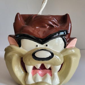 Pode incluir: Uma vela de cerâmica do Diabo da Tasmânia com um pavio branco. A vela apresenta uma cabeça castanha, olhos pretos e um rosto bege com boca rosa e dentes brancos. É um item de colecionador.