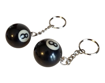 May include: Two black 8-ball keychains. Each keychain features a glossy black sphere with a white number 8. The spheres are attached to a silver chain and a keyring. The keychains are set against a white background.