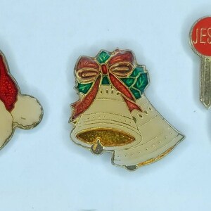 May include: Four Christmas-themed pins: a red and white Santa Claus face, a gold bell with a red bow and green holly, a red key with the word "JESUS" on it, and a red heart with the words "JESUS IS A FRIEND" in gold.