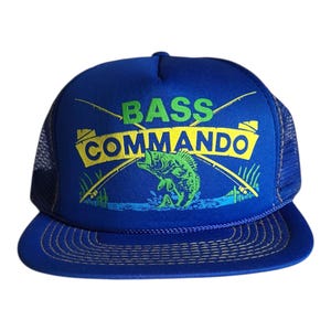 May include: A blue trucker hat with the words "BASS COMMANDO" in green and yellow. The hat features a graphic of a fish jumping out of the water, with fishing rods crossed behind it. The hat has a mesh back and a curved brim.