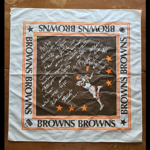 Puede incluir: Pa&ntilde;uelo cuadrado blanco y marr&oacute;n de los Browns con ribete y texto naranja. El centro presenta una ilustraci&oacute;n de un jugador de f&uacute;tbol americano y firmas. Las palabras "BROWNS" est&aacute;n impresas alrededor del borde. El pa&ntilde;uelo es de tela.