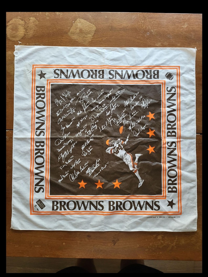 Puede incluir: Pa&ntilde;uelo cuadrado blanco y marr&oacute;n de los Browns con ribete y texto naranja. El centro presenta una ilustraci&oacute;n de un jugador de f&uacute;tbol americano y firmas. Las palabras "BROWNS" est&aacute;n impresas alrededor del borde. El pa&ntilde;uelo es de tela.