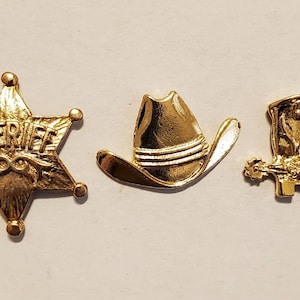 May include: Three gold-colored Western-themed charms. The charms include a sheriff's badge with the word "SHERIFF", a cowboy hat, and a cowboy boot with a spur. These charms are likely used for jewelry making or crafting.