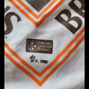 Puede incluir: Tela blanca con dise&ntilde;o geom&eacute;trico marr&oacute;n y naranja. La tela tiene las letras "BR" y "S" en marr&oacute;n. Se ve un logotipo de la NFL "Producto con licencia oficial", junto con el a&ntilde;o 1990.