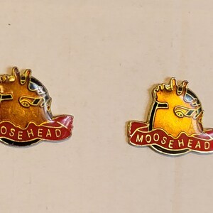 Op de afbeelding: Twee gouden spelden met een zwarte rand en een rood lint. Het lint heeft de tekst "MOOSEHEAD" in witte letters. De spelden tonen een gestileerd elzenhoofd in geel.
