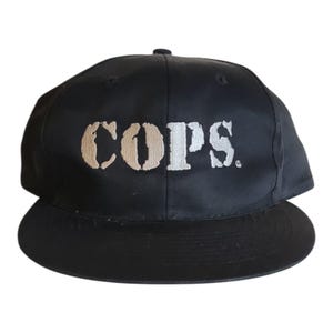 Puede incluir: Gorra de béisbol negra con visera plana. La palabra "COPS" está bordada en beige y blanco en la parte delantera. Un estilo de gorra clásico, adecuado para ropa informal. La gorra es de tela oscura.