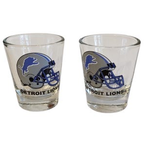 Puede incluir: Dos vasos de chupito transparentes con el logotipo de los Detroit Lions. El logotipo incluye un casco de fútbol azul con un león plateado y las palabras "DETROIT LIONS" en negro. Los vasos son de cristal transparente.