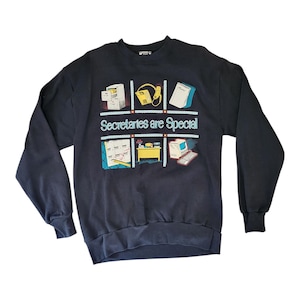 Pode incluir: Camisola azul marinho com um design gráfico colorido. O design apresenta imagens relacionadas com o escritório, como um arquivo, um telefone e um computador, dispostas em uma grade. O texto "Secretaries are Special" está impresso abaixo.