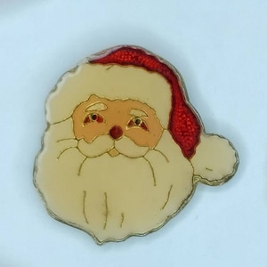May include: A red and white Santa Claus face ornament with a white beard and a red hat. The ornament is made of glass and has a shiny finish.