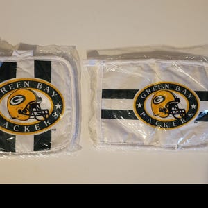 Op de afbeelding: Twee Green Bay Packers-items in plastic verpakking. Eén is vierkant met zwart-witte strepen en een teamlogo. De andere is ovaal van vorm met hetzelfde ontwerp. Beide hebben het logo van het team met een gele helm.