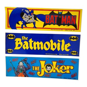 May include: Three vintage Batman stickers. The top sticker shows Batman with the text "BATMAN" in red and black on a yellow background. The middle sticker says "the Batmobile" in yellow on a blue background. The bottom sticker features the Joker with "THE JOKER" text.