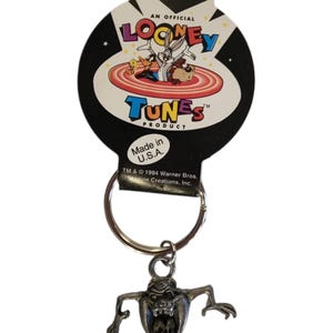 May include: A Looney Tunes keychain featuring the Tasmanian Devil character. The keychain has a silver-colored metal charm of the character and a metal keyring. The packaging reads "An Official Looney Tunes Product" and "Made in U.S.A."