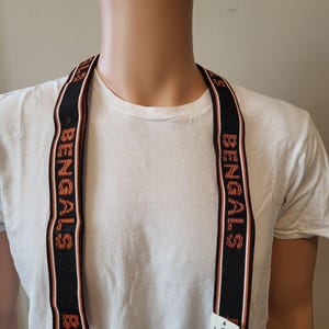 May include: Black suspenders with the word "BENGALS" in orange and white lettering. The suspenders have a black background with orange and white stripes. The suspenders are clipped to a white t-shirt.