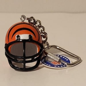 May include: Miniature orange and black football helmet keychain. The helmet has black stripes and a black facemask. A silver chain and key ring are attached. A small tag with text is also attached.