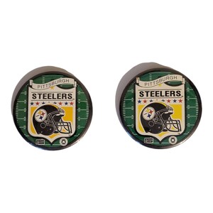 May include: Two circular Pittsburgh Steelers team logo pins. Each pin features the team's helmet logo on a yellow background, with the word "STEELERS" and "PITTSBURGH" above. The background is green with white yard lines.