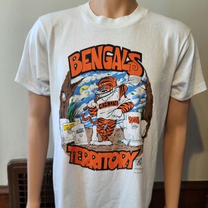 May include: White t-shirt featuring a cartoon tiger in a Cincinnati Bengals uniform, holding a shovel. The shirt has the words "Bengals Territory" in orange and black. The design includes tombstones and a tree.