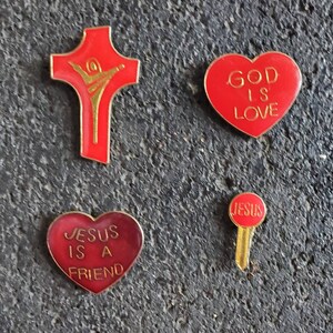 May include: Four red and gold pins with religious messages. The pins are shaped like a cross, a heart, and a key. The cross has a gold figure of a person on it. The heart pins say "God is Love" and "Jesus is a Friend". The key pin says "Jesus".